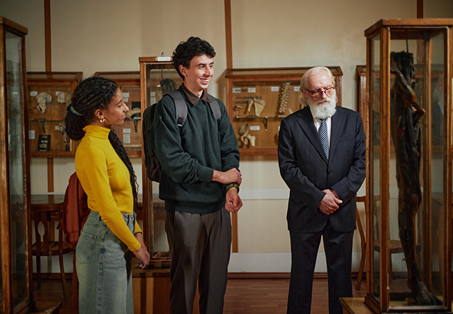 an older man and two students stand in a museum admiring a piece