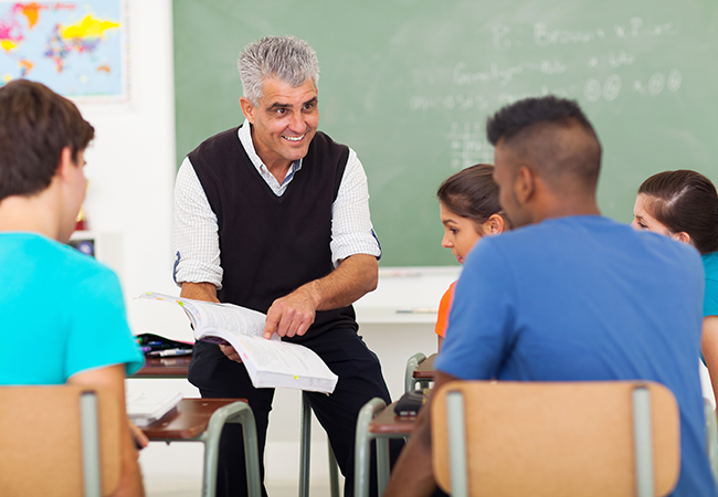 a teacher sits on top of a desk while pointing at a book showing his students