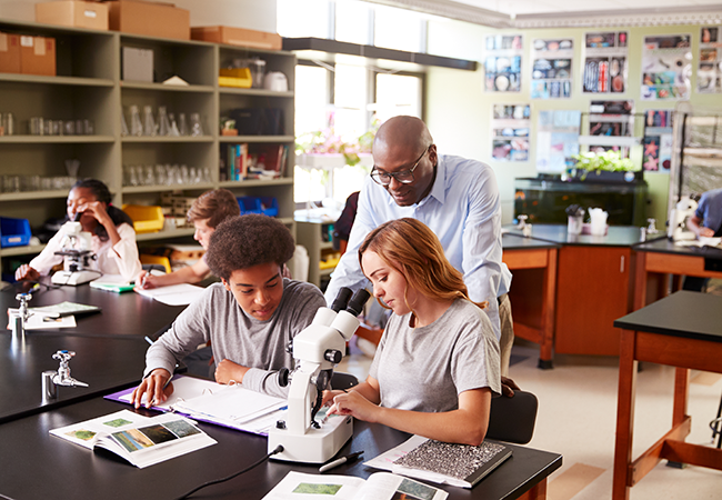 a teacher leans over students who are using a microscope