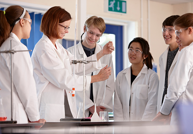 an instructor stands in the middle of students conducting an experiment with beakers and all wearing white coats