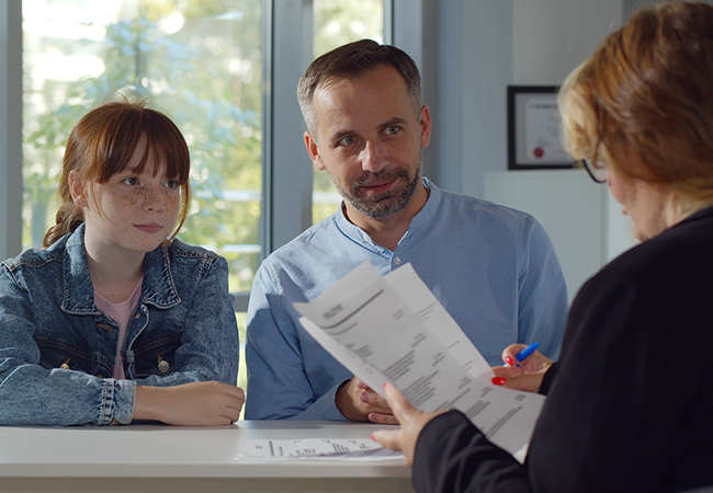 a father and daughter sit at a table across from a school admin during a meeting