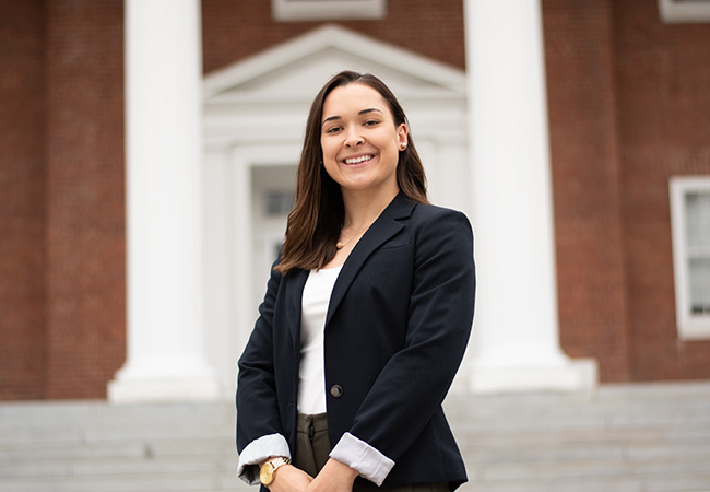 a young woman stands in front of college building smiling at camera