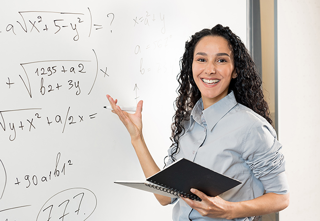 a female math teach smiles at camera while pointing at math equations on the board