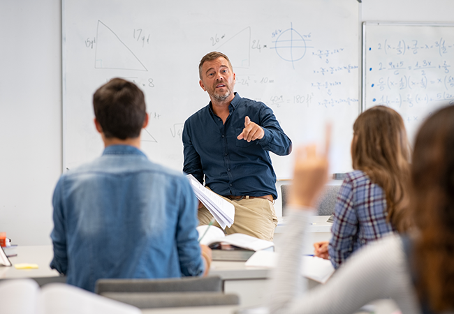 a teacher sits on his desk at the front of a class while a student rasies a hand