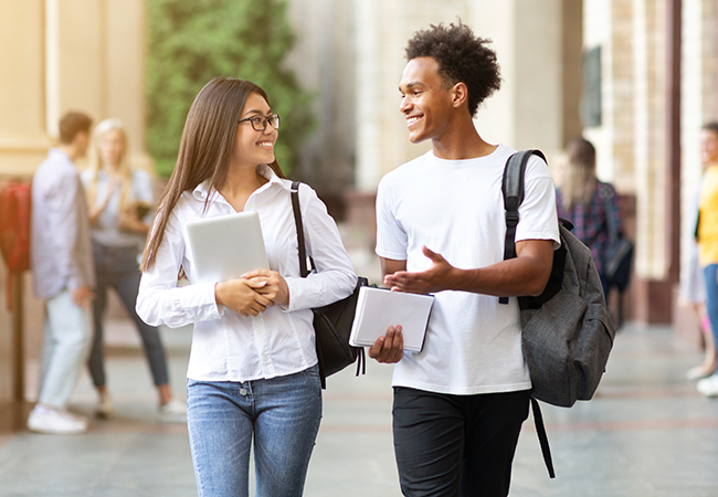 2 students holding backpacks and laptops walk on campus smiling at eachother