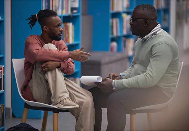 two men sit in chairs facing eachother talking