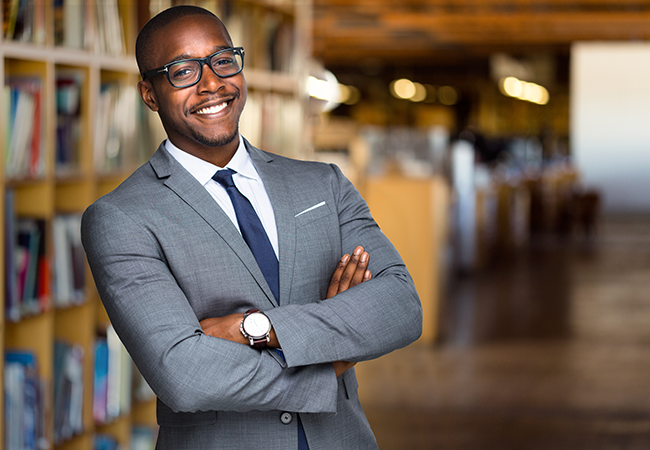 a man in a suit in a library crosses his arms and smiles towards camera
