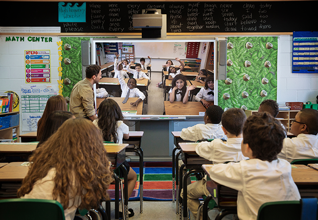 a classroom of young students video chats on a large screen with another classroom filled with young students