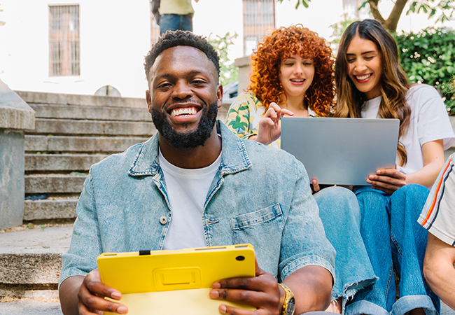a man sits with his ipad smiling at camera with two women behind him on a laptop
