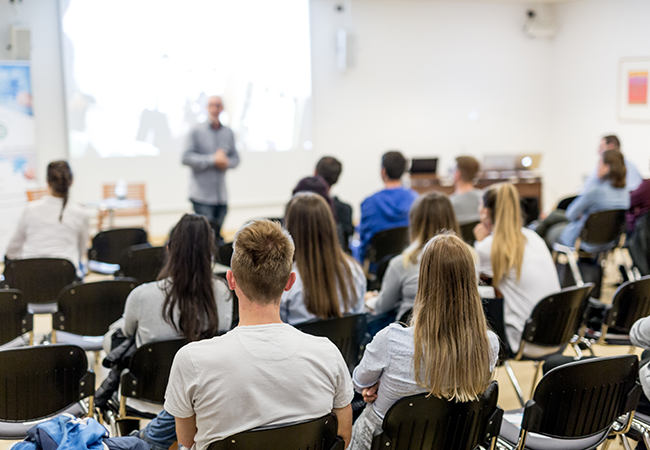 a wide shot of a very full classroom with the instructor at the front blurred out
