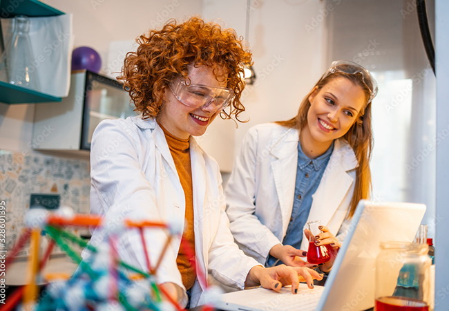 2 women in lab coats are working together and smiling in a lab with a laptop