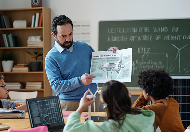 a teacher stands in front of students holding up a book and pointing at it