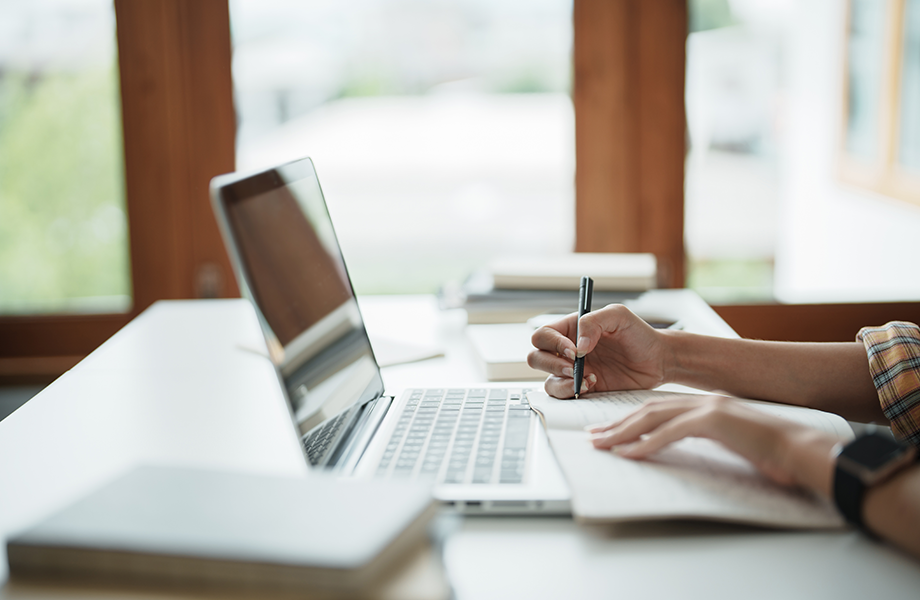 laptop on desk with person writing on paper next to the computer