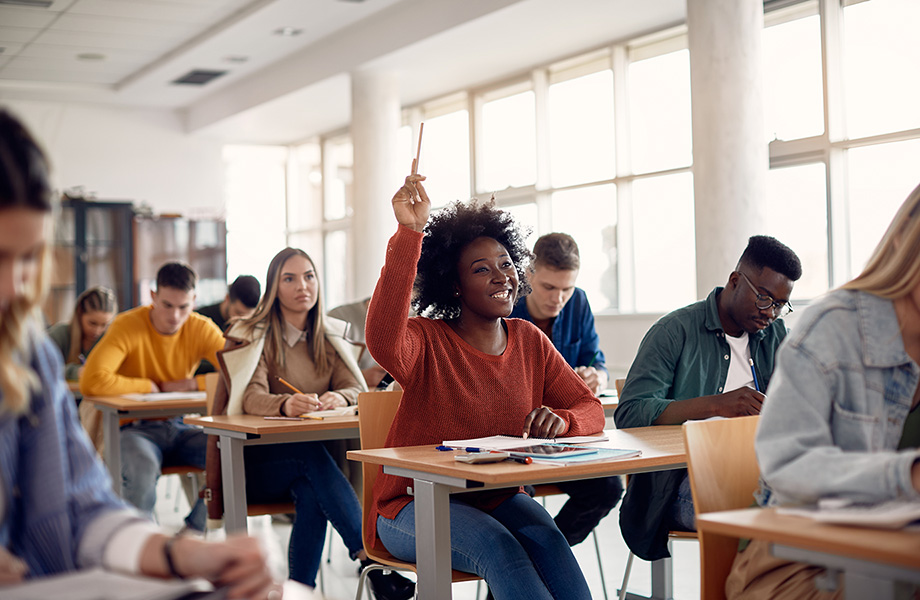 a student sits in a classroom with attention towards the front smiling and raising hand