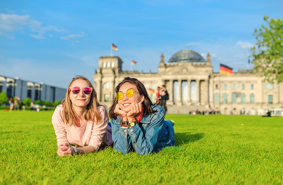 2 students lay on their stomaches in a grassy field smiling at camera in front of a large building with german flag in background