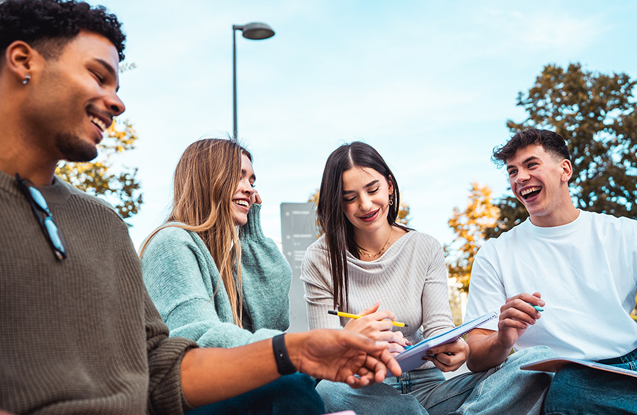 4 students sit on the grass with books and pens talking and laughing