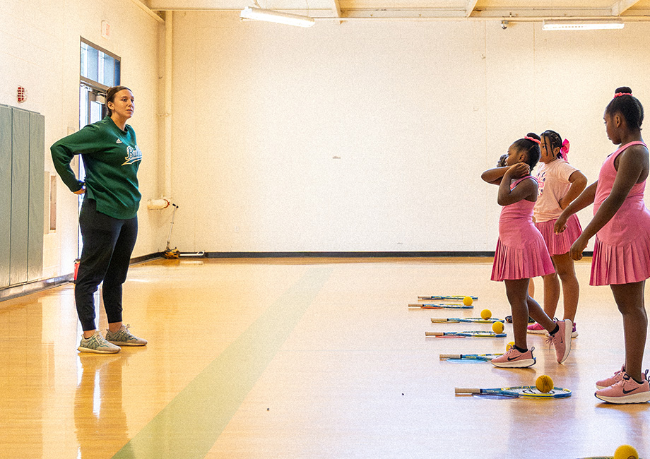 a female coach stands in front of children with tennis rackets