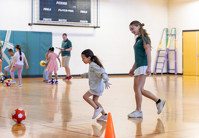 a woman coach follows a young girl smiling in gym kicking a soccer ball