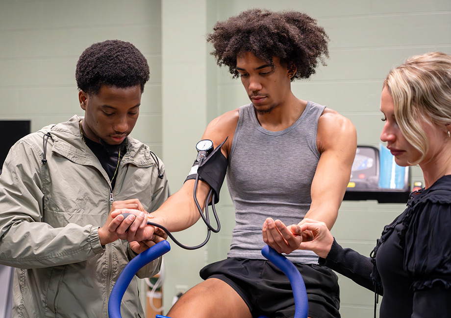 2 athletic trainers stand and take blood pressure from man on bicycle