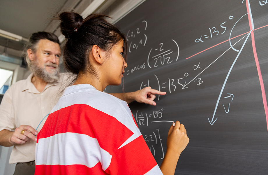 a teacher stands behind a student at the blackboard working on a math equation