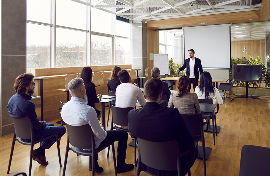 a man stands at the front of a room of professionals at work doing a presentation