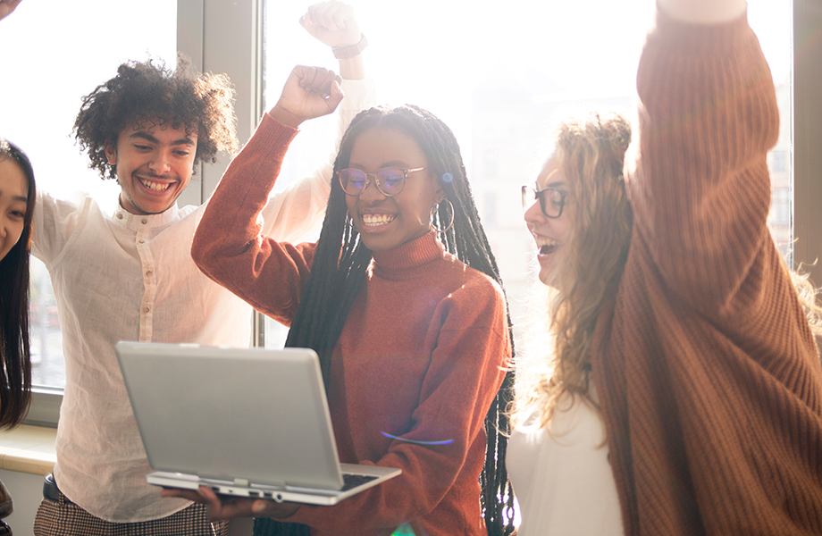 a group of older students surround a woman on a laptop holding hands up in the air celebrating