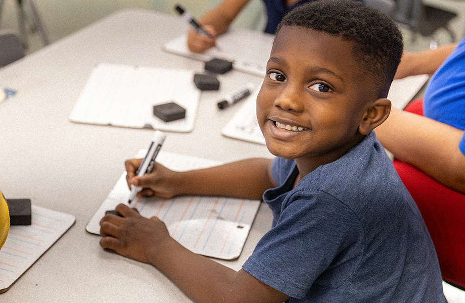 a young student smiles to camera while working on handwriting in a classroom