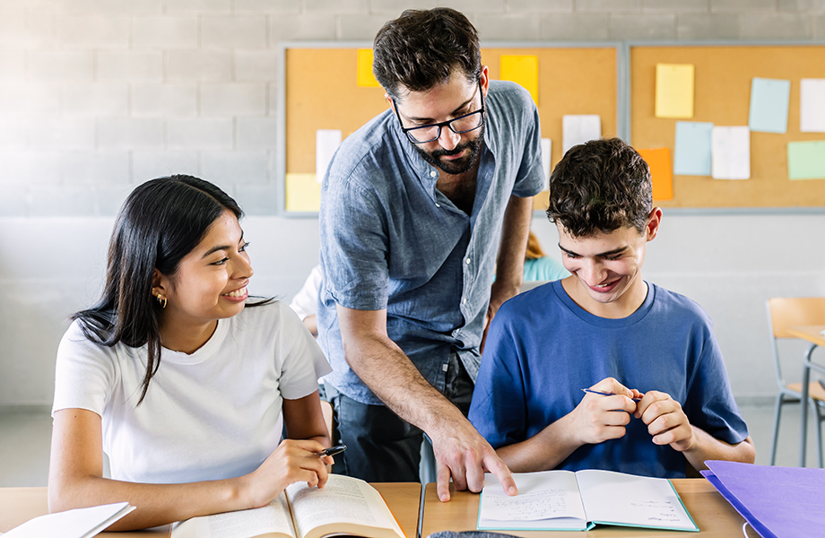 a male teacher leans over 2 students smiling with opens books on desk