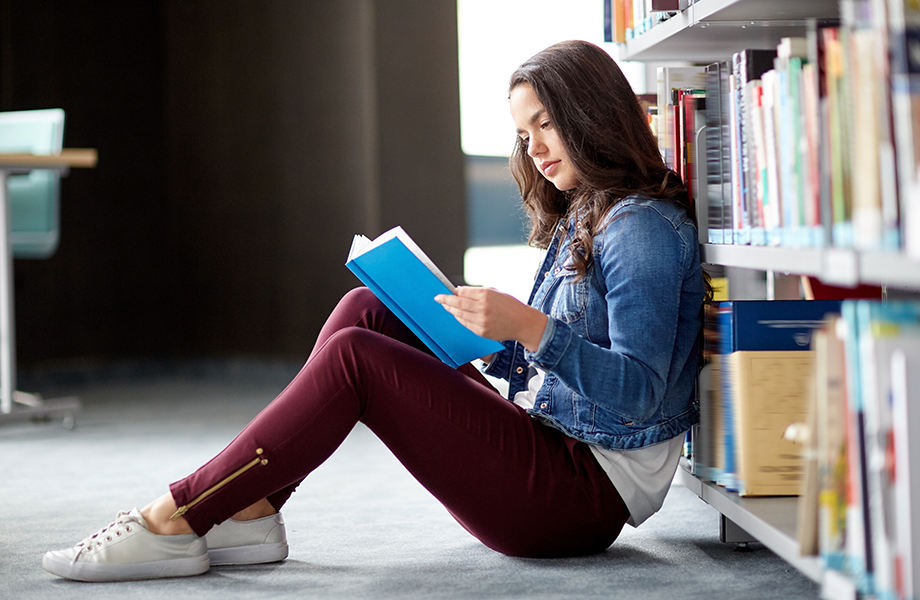a young high school girl sit and leans up against a rack of books reading a book in her library