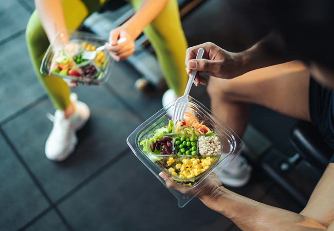 close up of two people in a gym sitting on bench eating healthy food