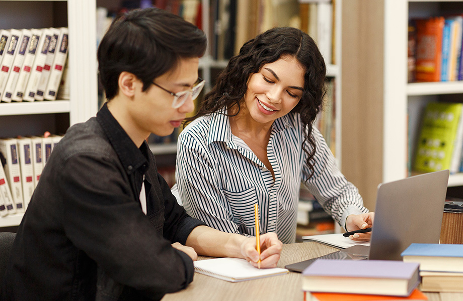 man and woman sitting at desk in libarary with books and laptop on table