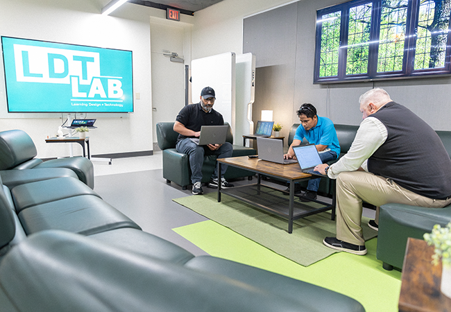 3 men sit at couches working on their laptops inside the learning and design teaching lab at usf. 