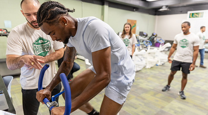 3 students look on as athlete uses stationary bike