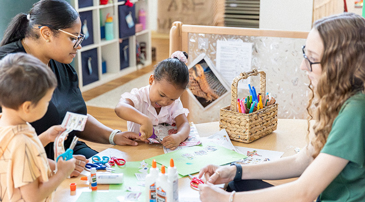 a few children and teachers sit at a table cutting and gluing paper
