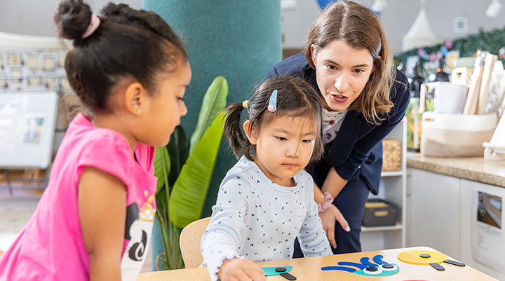 a teacher stands over 2 young girls at a table who are doing arts and crafts at table