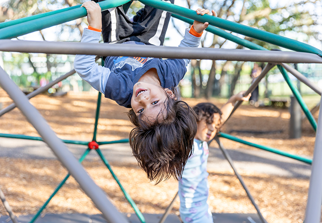 a young boy hangs upside down on a jungle gym smiling at the camera