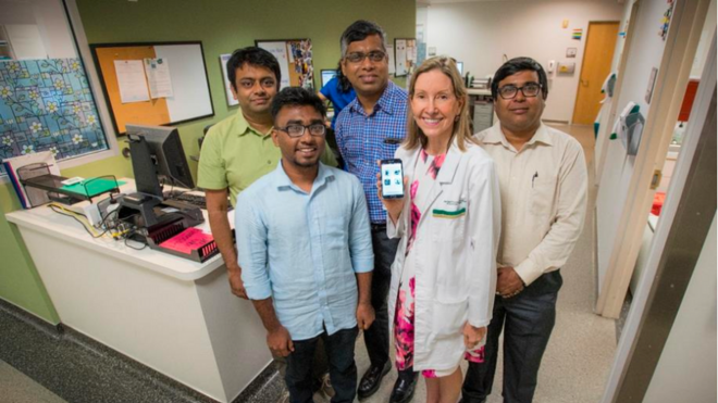 A group of five professionals, including a doctor in a white coat, pose together in a hospital or clinic setting. The doctor is holding up a smartphone displaying an application, suggesting a medical or healthcare-related tech development.