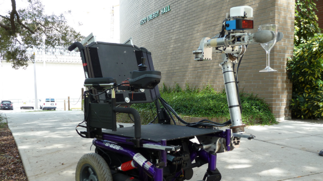  modified robotic wheelchair with a robotic arm is positioned outside a building labeled "ENG ENERGY HALL." The robotic arm is holding a wine glass, demonstrating its precision and assistive capabilities.