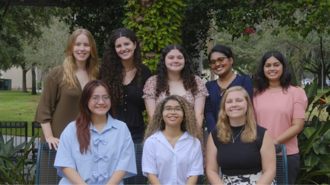 A group of eight people pose together outdoors in a lush, green setting, smiling at the camera. They are dressed in a mix of casual and semi-formal attire, suggesting a professional or academic gathering.