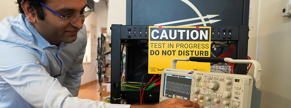 Professor turns knob on equipment, with "caution" sign in the background