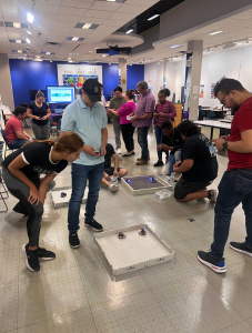 Participants test their assembled robotics kits in an interactive workshop setting, gathered around demonstration tables in a brightly lit room. The activity showcases collaborative problem-solving and applied learning in STEM.