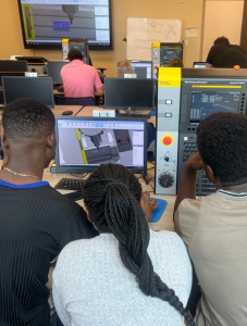 Students collaborate on a computer-aided manufacturing (CAM) project, working with specialized software and equipment in a classroom setting. The scene emphasizes teamwork and skill development in advanced technology applications.