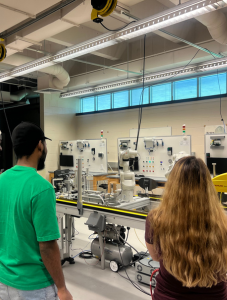 Two participants observe a robotic arm and conveyor system in a modern engineering lab, surrounded by industrial equipment and instructional displays. The setting highlights hands-on learning in automation and robotics.