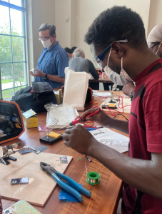 A participant carefully works on an electronics project using a soldering iron, surrounded by tools and components at a workshop. The activity emphasizes hands-on skill-building in electrical engineering and circuit design.