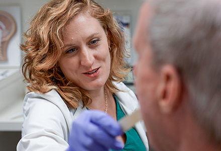 A woman with medium length wavy red and blonde hair using a wooden tongue depressor to treat an older male patient in a clinical exam room.