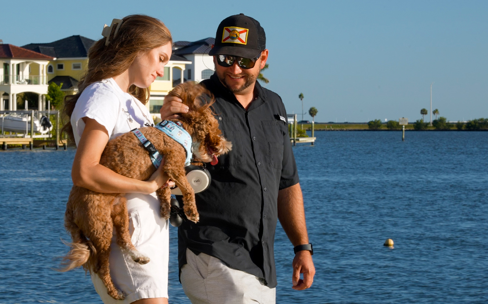A woman holds a small fluffy dog while a man pets him at sunset in ront of water.