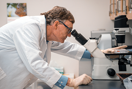 Dr. Stephen Liggett wears a white lab coat and glasses while looking through a microscope in his laboratory. Scientific equipment and materials are visible on the lab bench around him.