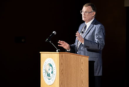 Dr. Stephen Liggett speaks at a wooden podium bearing the USF seal, wearing a blue blazer and lanyard. He gestures while speaking into a microphone against a dark background during a formal presentation.