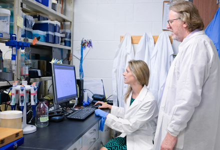 Two scientists discuss findings while wearing white lab coats and looking at a computer.