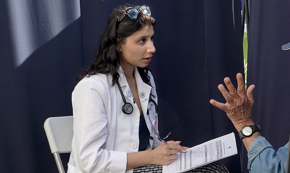 A young woman wearing a white medical coat and stethoscope sits in a folding chair, holding a clipboard and pen. She is looking intently at a person seated across from her, whose hand is raised in conversation. The setting appears to be an outdoor community health event.